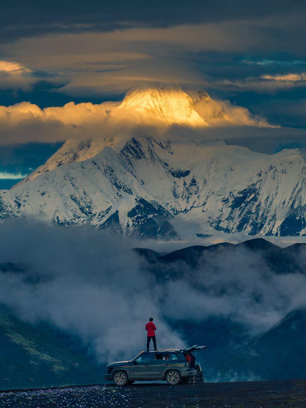 Traveler on off-road vehicle roof overlooking snow mountains, Western Sichuan